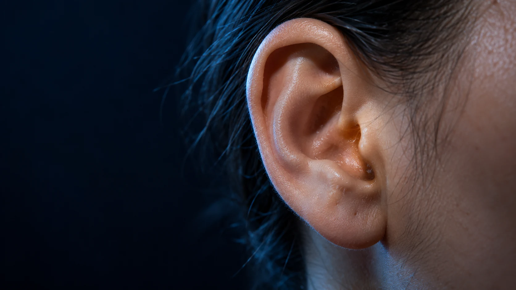 Close view of a healthy human outer ear against a dark navy background with subtle blue and amber light